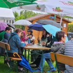 Gäste auf Bierbänken mit aufgespannten Regenschirmen